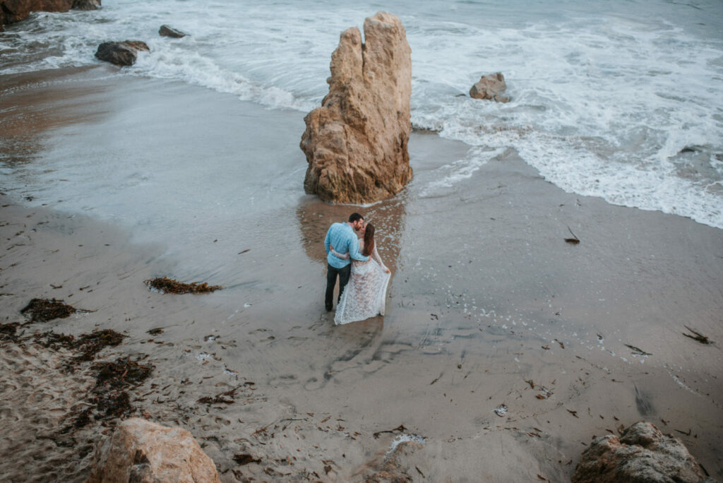 Follow the beautiful waves from the top view of El Matador BEach, Los Angeles where a pregnant couple poses by the big rock right on the shore during their maternity photography session with Dear Birth