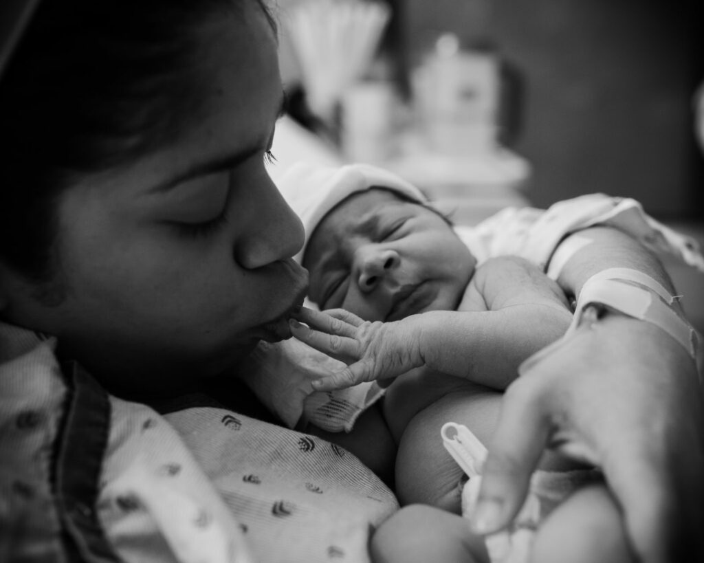 black and white image of mom close up kissing her newborn daughter tiny fingers,a s she sleeps peacefully in her arms right after birth. Photography by Dear Birth for Olivia's birth story