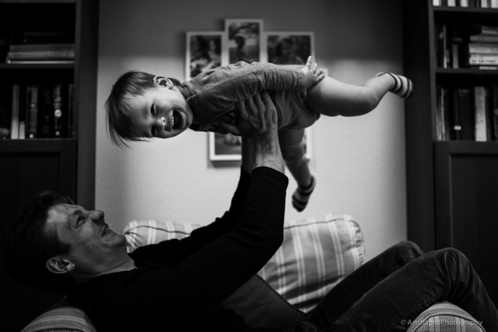 Black and white image of father holding his son in the air while he laughs and enjoys the ride looking at the camera of Dear Birth in Santa Monica, CA.