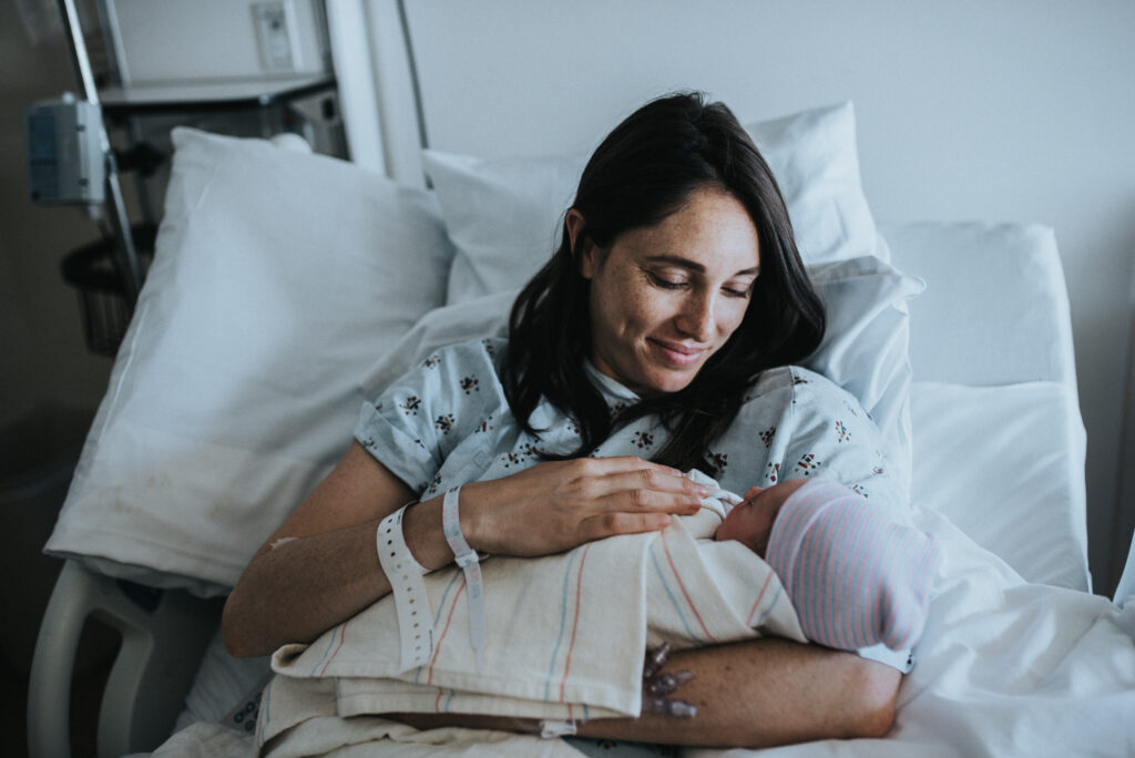 Color image of when big brother meets his newborn baby brother. Mother holding a newborn baby wrapped in a hospital blanket and hat. Mother wears a hospital gown and looks down at her baby. Image by Los Angeles Birth Photographer Diana Hinek for #dearbirth