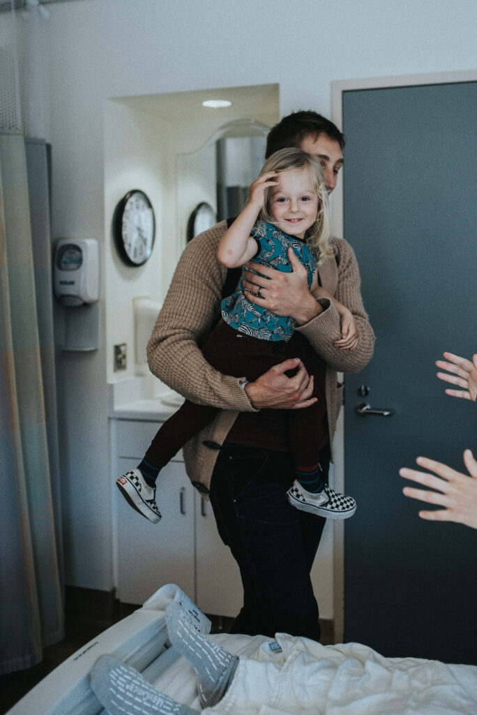 Color image of a man holding a toddler in his arms. Child looks at the camera while moving his hair from his face. Two hands out of frame reach out toward the child. Image by Los Angeles BIrth Photographer Diana Hinek for #dearbirth.