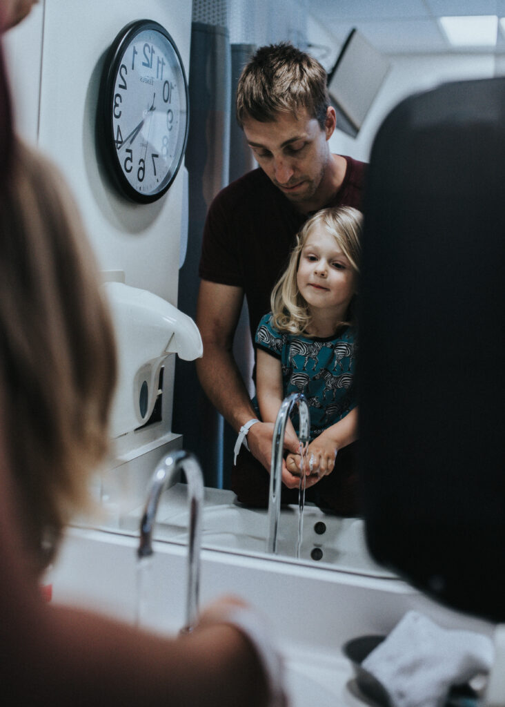 Color image of man washing the hands of a toddler in front of a mirror. A clock is visible on the wall next to them. Child is looking at the mirror. Image by Los Angeles birth photographer Diana Hinek for #dearbirth