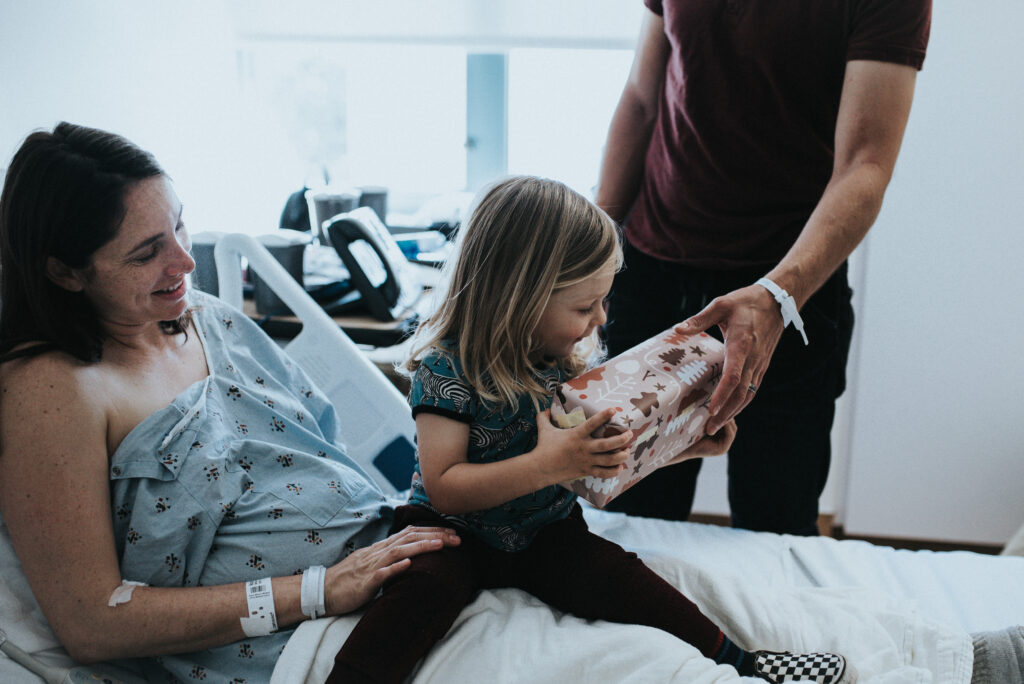 Color image of toddler sitting on the bed in a hospital bed where his mother is laying wearing a hospital gown. The child receives a wrapped up gift from the hands of a man standing next to them. Image by Los Angeles birth photographer Diana Hinek for #dearbirth.
