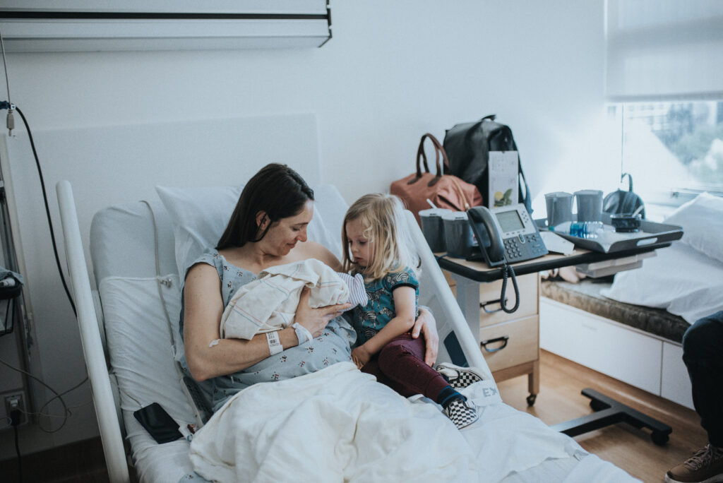 Color image of a mother in a hospital bed holding a newborn baby. Next to her is an older child looking at the baby. Image by Los Angeles birth photographer Diana Hinek for #dearbirth