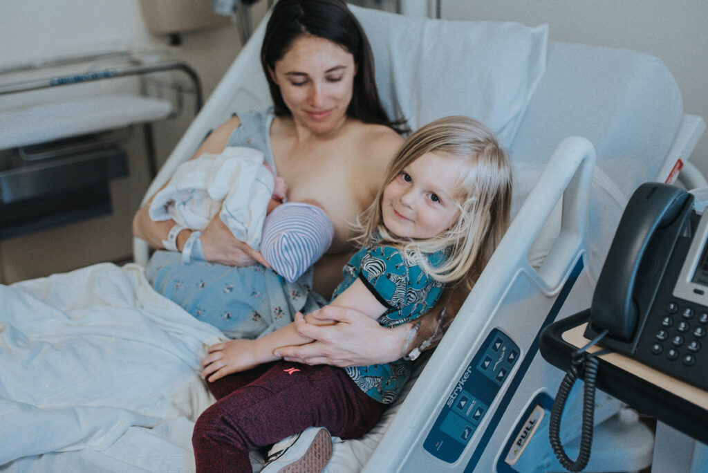 Color image of toddler sitting in a hospital bed with his mother who is hugging him and also holding a newborn to her breast. The older child is looking toward the camera. Image by Los Angeles birth Photographer Diana Hinek for #dearbirth.