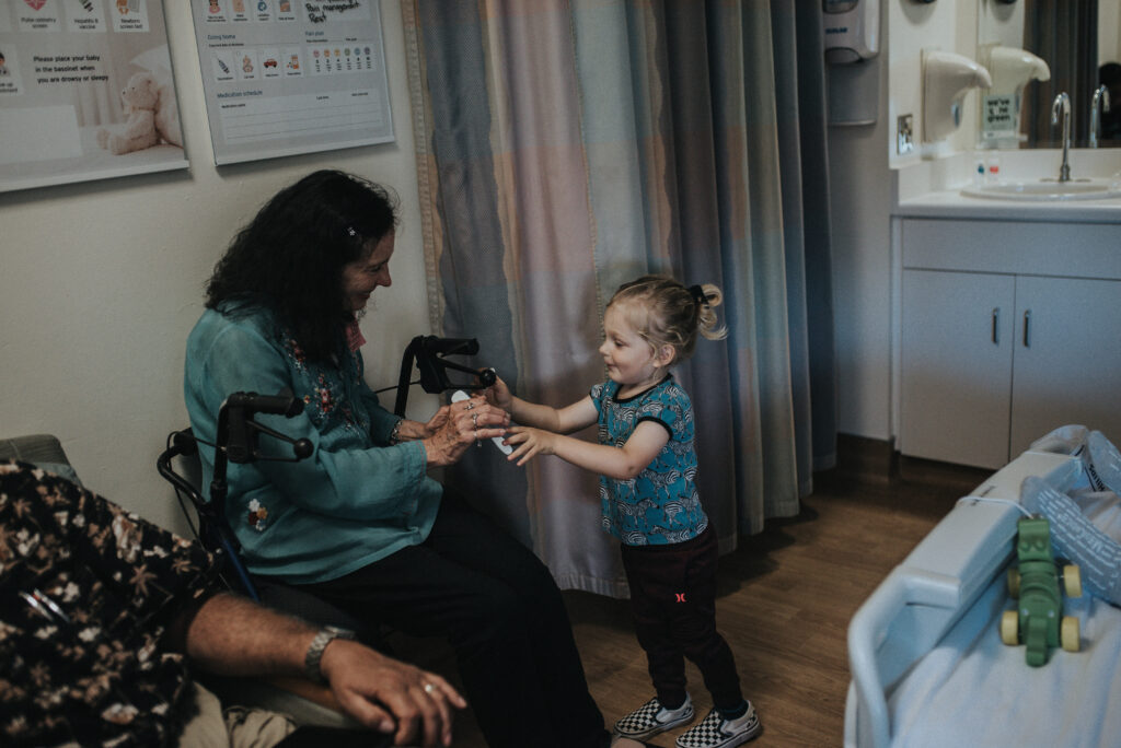 Color image of woman sitting on a wheelchair and reaching out to a toddler who is standing next to her. Thye are in a hospital room. Arms of another person come into the frame. Image by Los Angeles birth photographer Diana Hinek for #dearbirth.