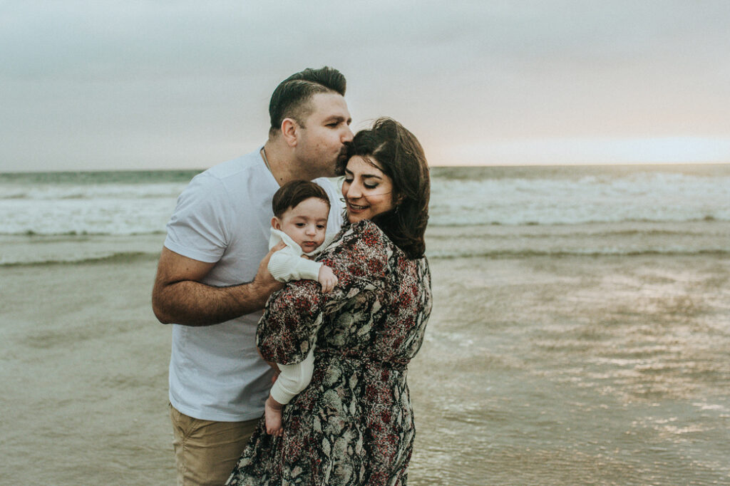 Color image of family of three kissing at the beach on a winter day in Santa Monica during a spectacular family session on a stormy day as photographed by Los Angeles Santa Monica family Photographer Diana Hinek