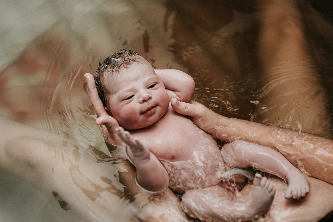 Baby looks up at her mom with her hand risen right after a home water birth and captured by Los Angeles birth photographer and videographer Diana Hinek for Dear Birth