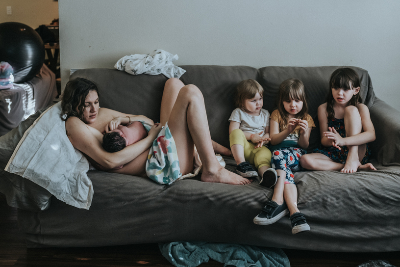mother sits on her couch holding her newborn baby while her other 3 girls chat next to her as captured byt Los Angeles birth photographer and videographer Diana Hinek for Dear Birth